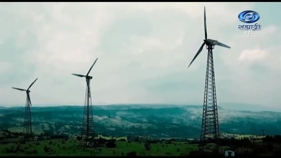 Three wind turbines stand tall against a cloudy sky, their blades slowly turning.  The scene is broadcast by DD Sahyadri, showcasing the green energy initiatives in India.

