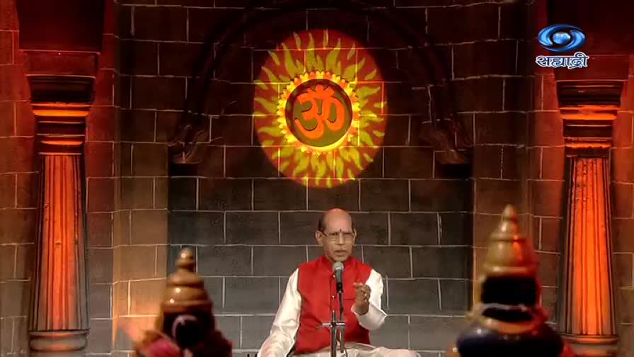 A man in a red vest speaks into a microphone, gesturing with his right hand. Behind him, a glowing symbol projects onto a brick wall, the DD Sahyadri logo visible in the corner.
