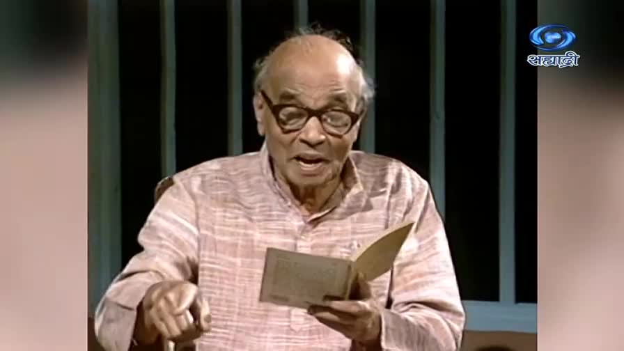 An elderly man with glasses is reading from a small booklet, his mouth open as he speaks. The DD Sahyadri logo is visible in the corner.
