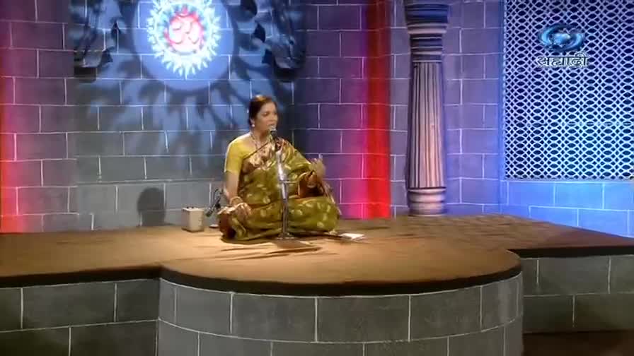 A woman in a patterned sari sits cross-legged on a stage, speaking into a microphone. The DD Sahyadri logo is visible in the background, and a spotlight shines on the performer.
