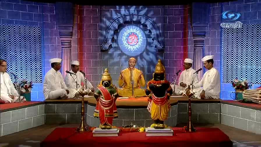 A man in a yellow robe sits center stage, speaking into a microphone. Two elaborately dressed statues flank him, while musicians in white outfits sit on either side, accompanying him.

