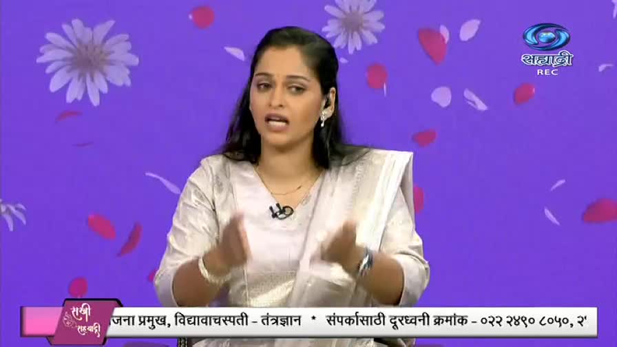 A woman in a light-colored sari gestures with her hands, speaking animatedly on a DD Sahyadri broadcast. The background is a vibrant purple, decorated with floating flower petals and a logo.

