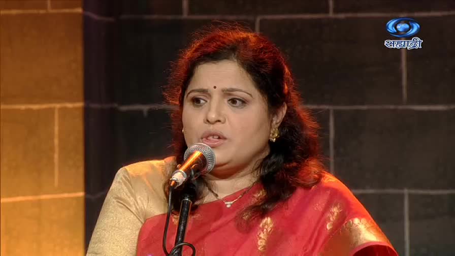 A woman in a red sari stands before a microphone, her mouth open as if singing. The DD Sahyadri logo is visible in the background.
