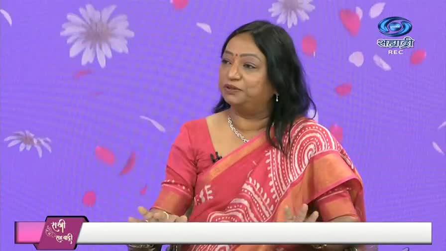 A woman in a red sari gestures with her hands, speaking on a television set. The backdrop is a soft purple with floating flower graphics and the DD Sahyadri logo.
