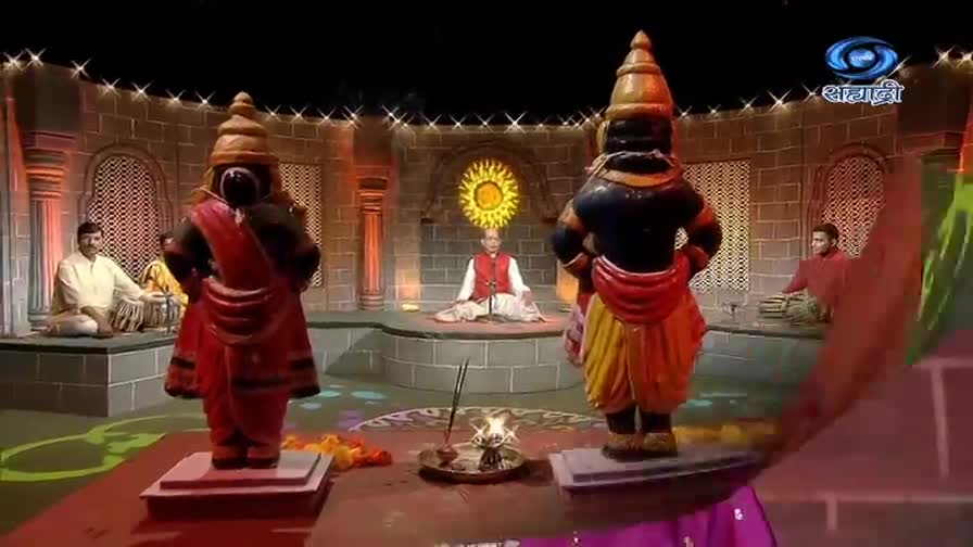 Two large statues, brightly colored, stand on pedestals, facing the audience. A man in a red vest sits cross-legged, speaking in front of a sun-like symbol, while musicians play nearby.
