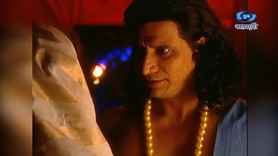 The man in the frame stares intensely, his expression fixed. A string of pearls hangs against his chest, and a white and gold garment is draped over his shoulder.
