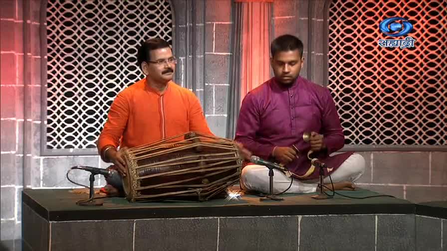 The man in orange strikes the barrel drum with his hands, while the other in purple keeps time with small cymbals. They're on a stage, likely for a DD Sahyadri broadcast.
