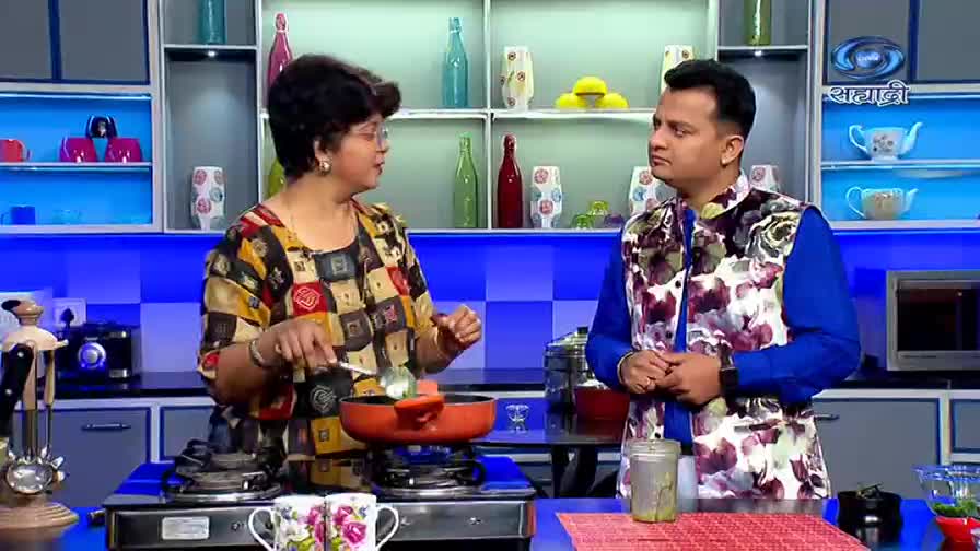 The woman in the patterned top stirs something in a small orange pan over a gas burner. The man in the floral vest looks on, listening intently.
