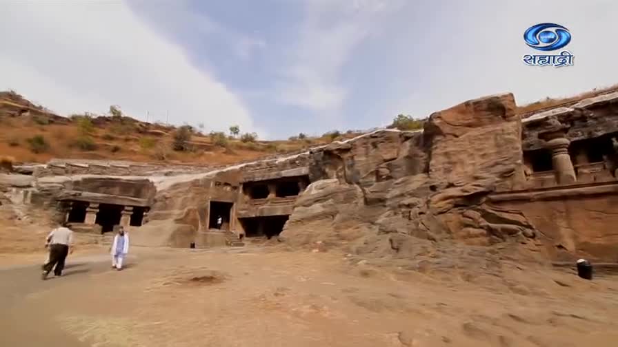 Two men walk across the open space in front of the ancient rock-cut architecture. The DD Sahyadri logo sits in the top right corner, above the weathered stone structures.

