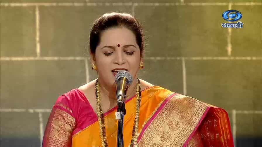 A woman in a vibrant saree sings into a microphone, her eyes closed in concentration. The DD Sahyadri logo is visible in the corner, suggesting a live performance.
