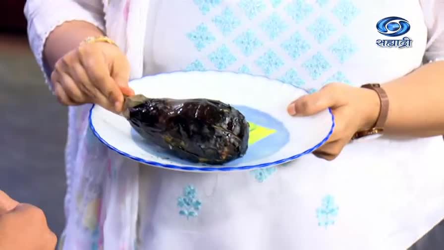 A woman, wearing a white kurta, holds a plate with a charred eggplant. She presents it for the camera on DD Sahyadri.
