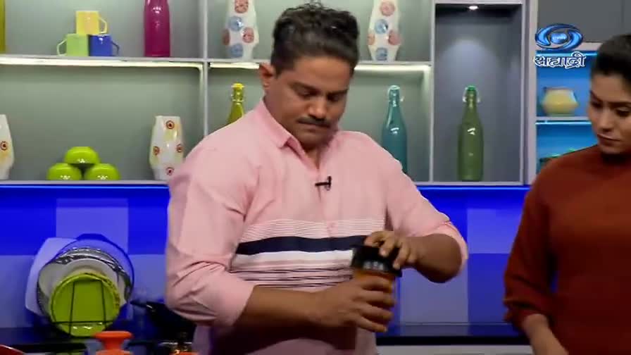 A man in a pink shirt is twisting the lid off a small container. The DD Sahyadri logo is visible in the background, likely a cooking show.
