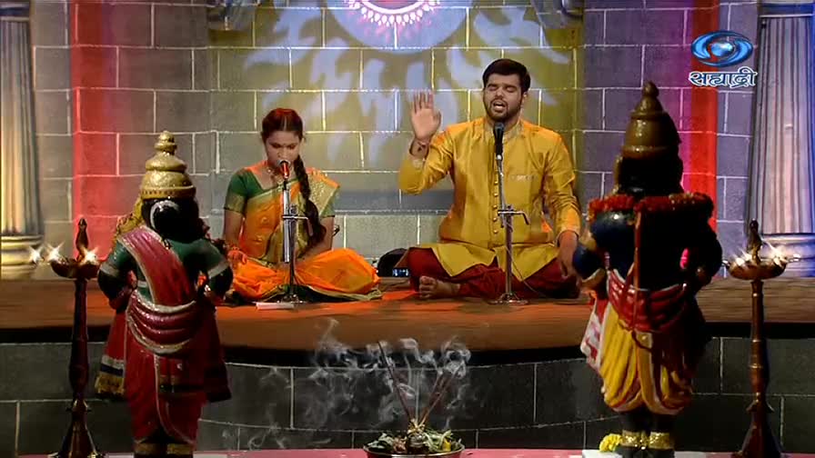 Incense smoke curls upward from a small container as a man in a yellow kurta raises his hand. A woman beside him, in a sari, sings into a microphone, while statues of deities flank them on either side.
