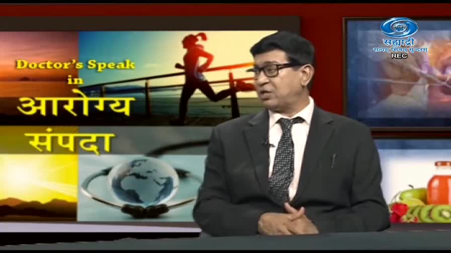 A man in a suit and tie is speaking, framed by the set of a show called "Doctor's Speak in Aarogya Sampada" on DD Sahyadri. The backdrop includes images of a runner, a stethoscope, and the channel's logo.

