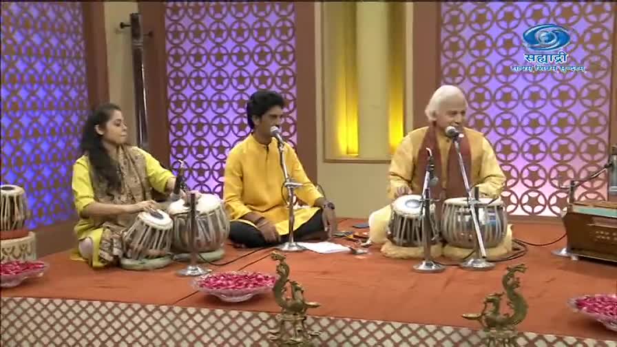Three musicians, each in a yellow kurta, are seated on a stage, playing instruments. The tabla players' hands move quickly, striking the drums in a rhythmic pattern.
