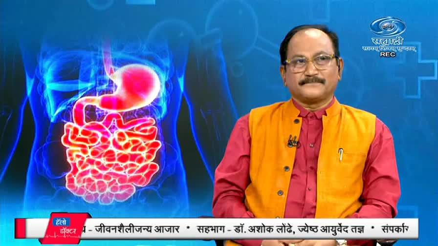 A man in a red shirt and orange vest is seated, speaking on a DD Sahyadri program. Behind him, a stylized diagram of the digestive system glows against a blue background.
