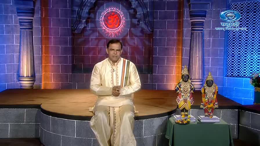 A man in cream-colored clothing sits on a raised platform, speaking directly to the camera. Behind him, two ornate statues stand on a small table, lit by the studio lights of DD Sahyadri.
