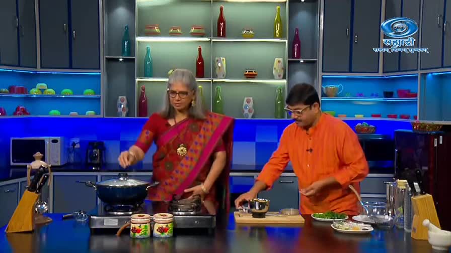 A woman in a red sari lifts the lid on a pot, while a man in an orange shirt works with a small grinder. They're in a brightly lit kitchen, likely on a DD Sahyadri cooking show.
