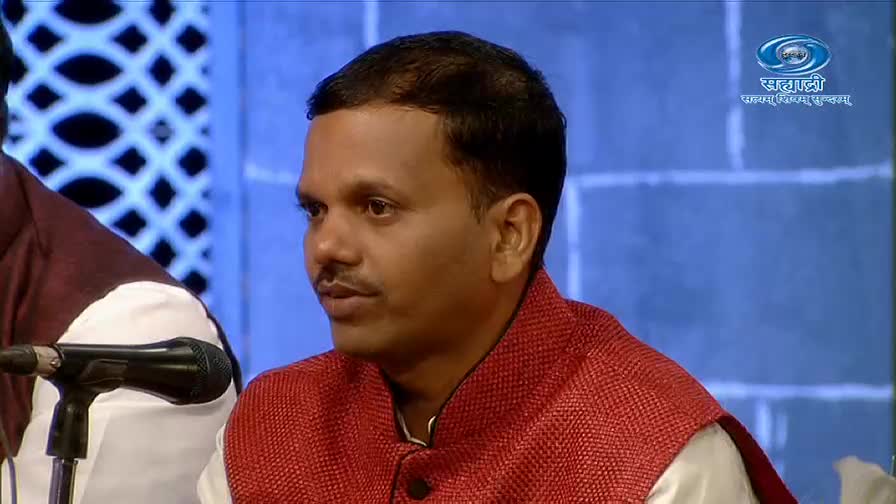 A man in a red vest sits, listening intently, near a microphone. The DD Sahyadri logo is visible in the background, suggesting a television studio setting.
