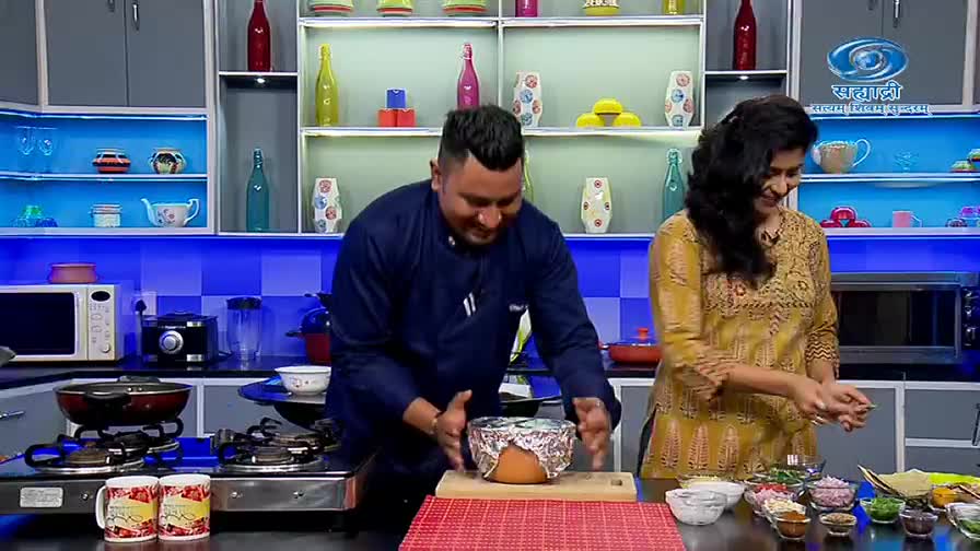 The chef, grinning, sets a foil-covered pot onto a wooden cutting board. A woman in a yellow kurta stands beside him, smiling as she prepares ingredients on the counter, likely for a DD Sahyadri cooking show.
