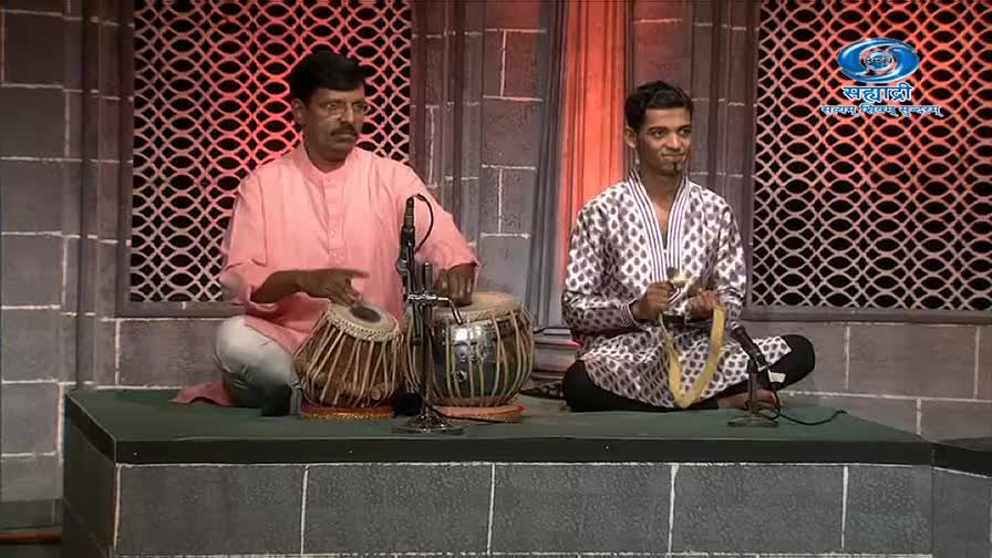 The man in the pink shirt is seated, hands moving quickly over the tabla drums. Beside him, the other musician holds a stringed instrument, ready to play.
