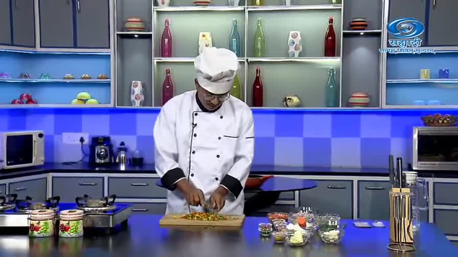 A chef in a white uniform expertly chops vegetables on a wooden board. The kitchen set, with its blue and gray tones, suggests a cooking show on DD Sahyadri.
