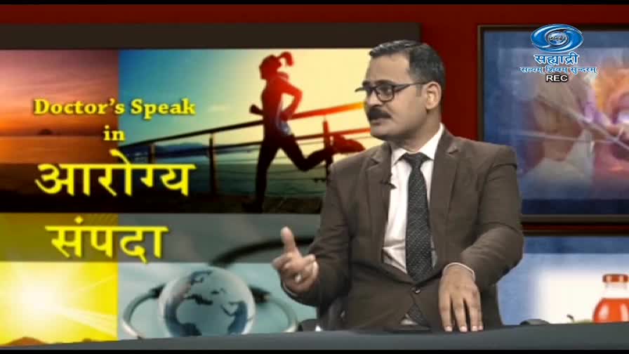 A man in a suit gestures with his hand as he speaks on a DD Sahyadri television show. Behind him, a screen displays the show's title in Marathi, with an image of a person running.

