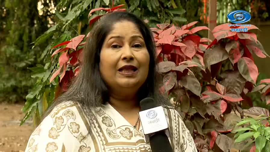 A woman speaks into a microphone, her patterned kurta a stark contrast to the deep red leaves behind her. The DD Sahyadri logo is visible on the microphone, indicating a broadcast from India.