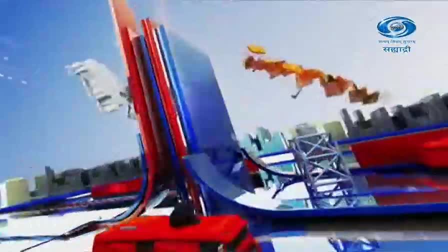 A red vehicle with a black roof sits on the ground. Above it, a large, abstract structure of red and blue panels rises towards a bright sky.