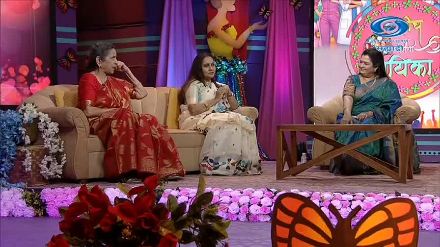 Three women sit on a sofa and chairs on a brightly decorated stage. The woman on the far left wears a red sari and rests her hand on her cheek. The woman in the middle is in a white outfit with floral accents. The woman on the right, dressed in a green sari, smiles as she looks towards the other two.