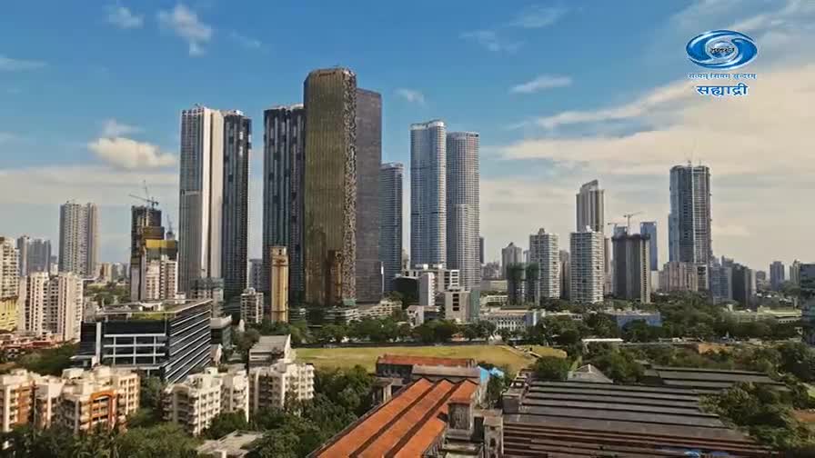The sky over Mumbai is a bright blue, dotted with a few white clouds. Tall buildings, some still under construction, stretch towards the heavens, their glass facades reflecting the sunlight. Below, a green field lies nestled amongst the urban landscape, a splash of nature in this bustling Indian metropolis.