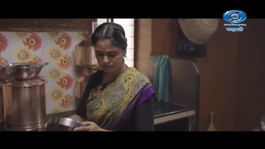 A woman in a sari carefully holds a metal bowl. She's in a kitchen, with copper pots and a floral-patterned backsplash behind her.
