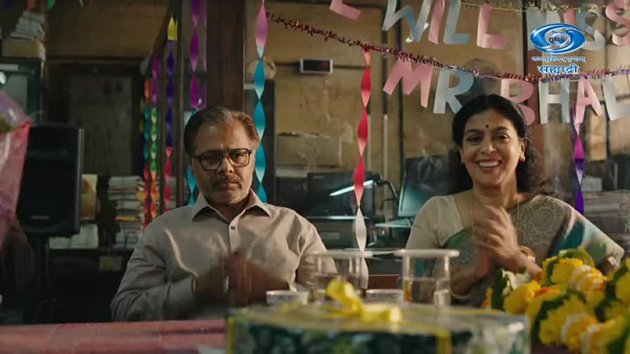 A man with glasses sits with a somber expression, while a woman beside him beams and claps her hands. Festive paper chains hang overhead, and a banner reads "WILL MISS MR BHA". A man with glasses sits with a somber expression, while a woman beside him beams and claps her hands. Festive paper chains hang overhead, and a banner reads "WILL MISS MR BHA".