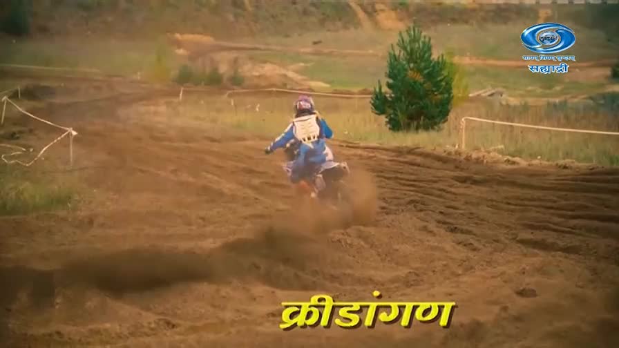 A dirt bike rider in blue gear kicks up a cloud of dust as they round a corner on the track. The DD Sahyadri logo is visible in the background. A dirt bike rider in blue gear kicks up a cloud of dust as they round a corner on the track. The DD Sahyadri logo is visible in the background.