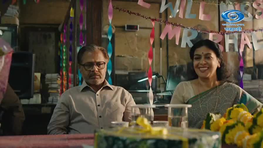 A man with glasses sits with a somber expression, while a woman beside him beams with joy. Festive paper streamers and a banner that reads "WILL MISS MR BHAI" hang behind them, suggesting a farewell celebration.