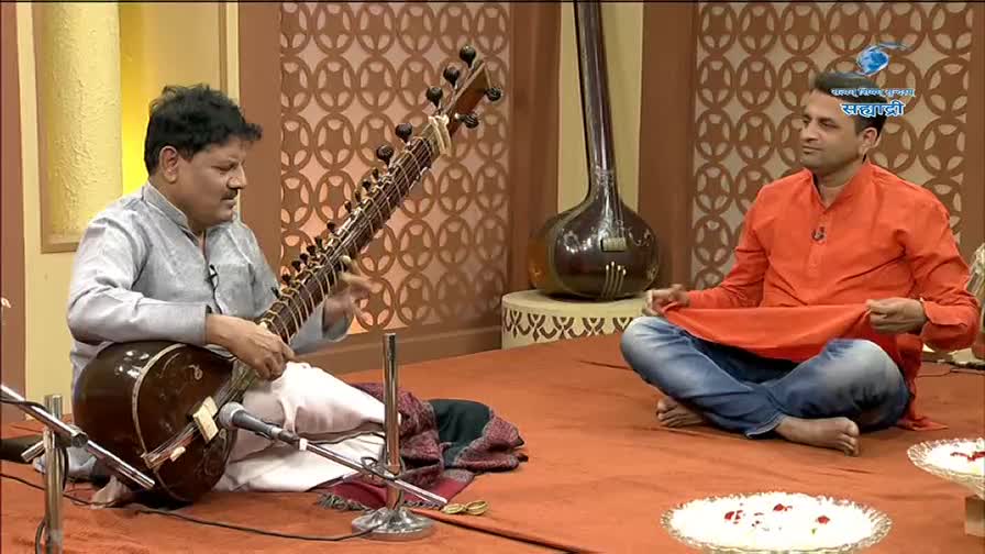A man in a grey kurta plays the sitar, his fingers deftly moving across the strings. Another man, dressed in an orange kurta and jeans, sits opposite him, holding a piece of fabric. A man in a grey kurta plays the sitar, his fingers deftly moving across the strings. Another man, dressed in an orange kurta and jeans, sits opposite him, holding a piece of fabric.