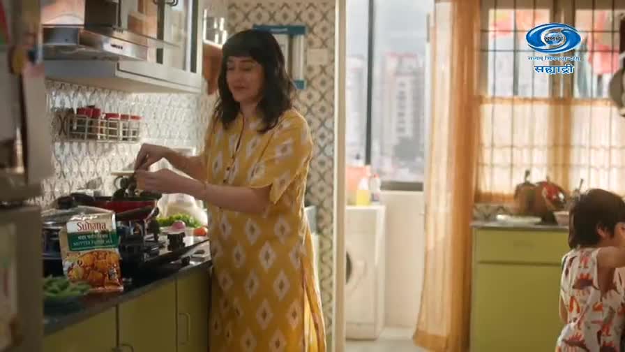 A woman in a yellow kurta stirs something in a pan on the stove. Her young son, wearing a dinosaur-print shirt, stands nearby. A woman in a yellow kurta stirs something in a pan on the stove. Her young son, wearing a dinosaur-print shirt, stands nearby.
