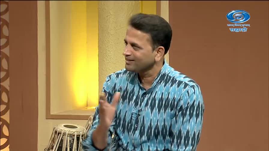 A man in a blue and black patterned shirt gestures with his hand while speaking. Beside him, a tabla rests on the floor, ready for a performance on DD Sahyadri.