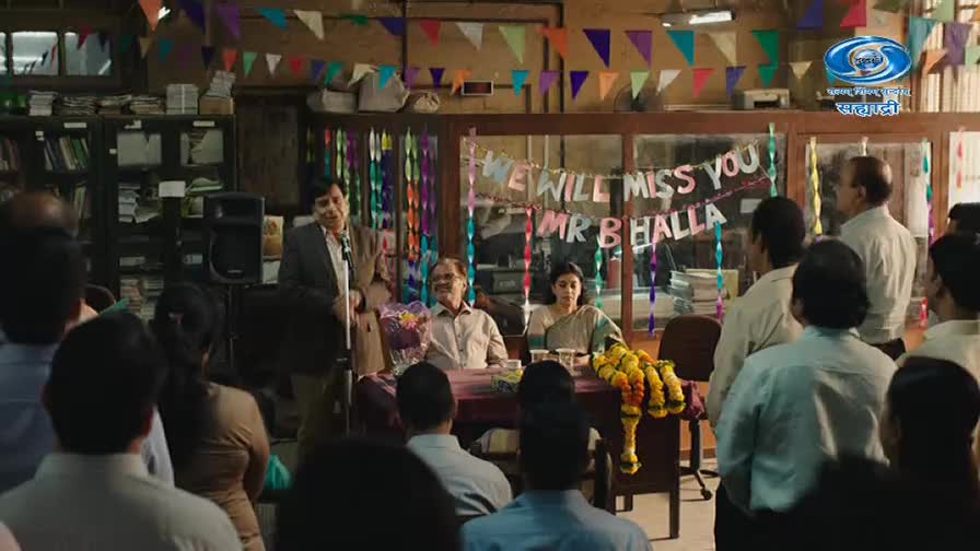 A man stands at a microphone, addressing a crowd gathered in a room decorated with colorful streamers and a banner that reads "We Will Miss You Mr B Halla." A couple sits at a table adorned with a garland of marigolds, likely the guests of honor.