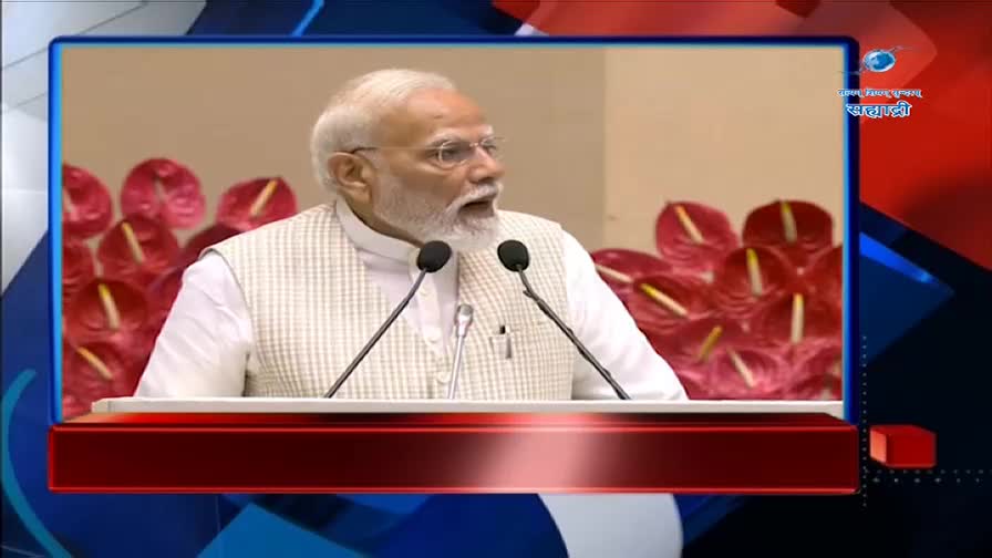 The Prime Minister speaks at a podium, his voice amplified by microphones. Behind him, a cluster of deep red anthurium flowers adds a splash of color to the backdrop.