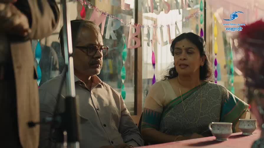 A man with glasses and a woman in a sari sit at a table, looking towards something out of frame. Colorful paper decorations hang behind them, hinting at a celebration.