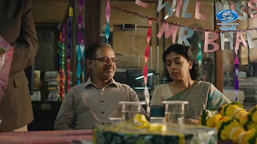 A man with glasses smiles as a woman in a sari looks down. Colorful streamers hang above them, spelling out "WILL MISS MR BHAI" for a DD Sahyadri broadcast.