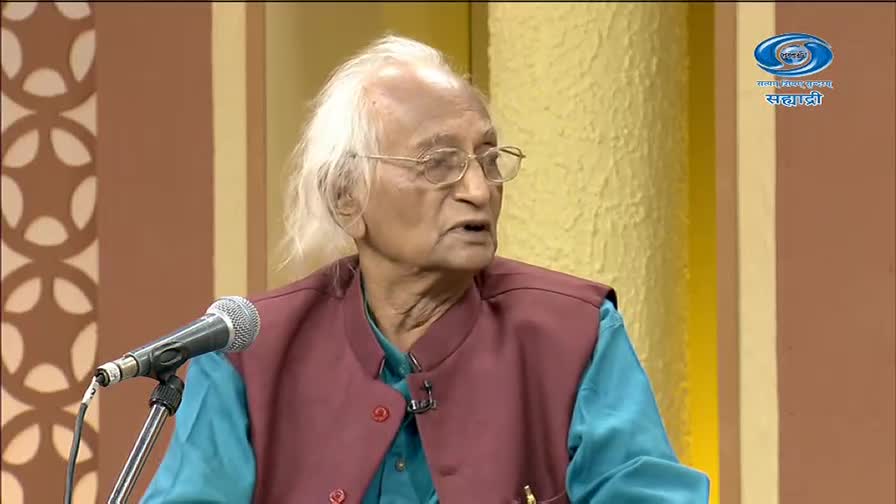 An older gentleman with white hair and glasses speaks into a microphone. He wears a teal kurta and a maroon vest, likely for a program on DD Sahyadri.