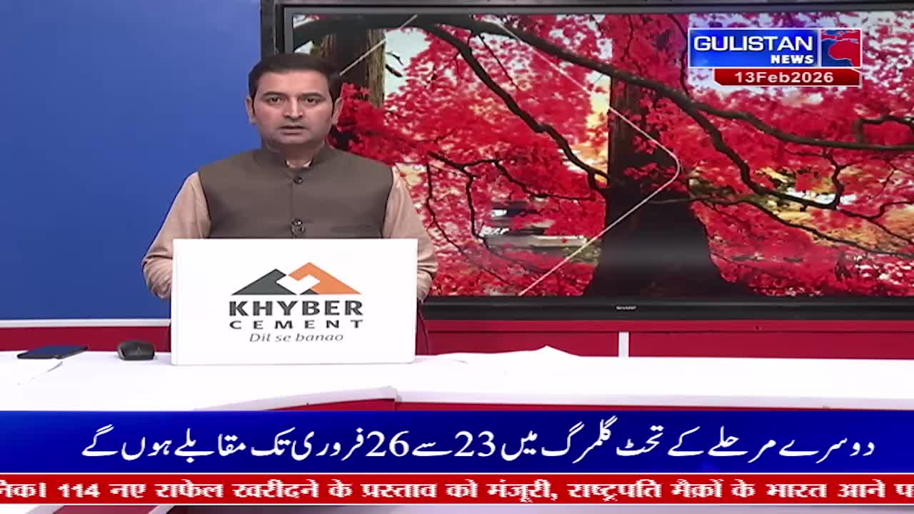 A man stands in front of a screen displaying vibrant red foliage and the Gulistan News logo. A white board with the Khyber Cement logo sits on a desk before him.