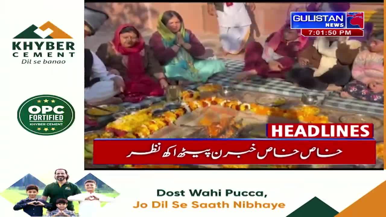 People are gathered, some with hands pressed together, around a spread of flowers and offerings. Gulistan News is broadcasting headlines about this event.