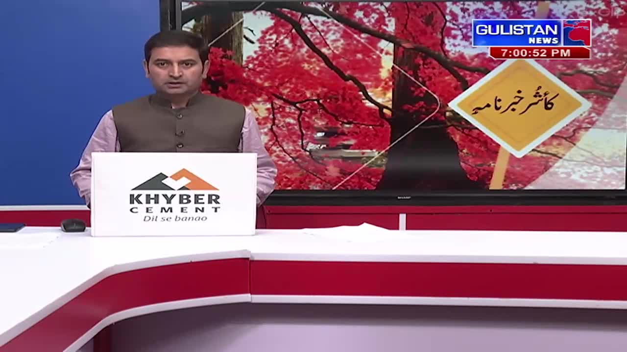 A man stands at a news desk, holding a sign for Khyber Cement. Behind him, the Gulistan News logo appears on a screen displaying a vibrant autumn scene and a yellow diamond-shaped sign with Urdu script.