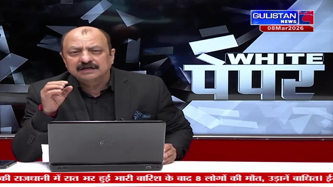 A man with a mustache gestures with his right hand while speaking. A laptop sits open before him, and a news chyron scrolls across the bottom of the screen.