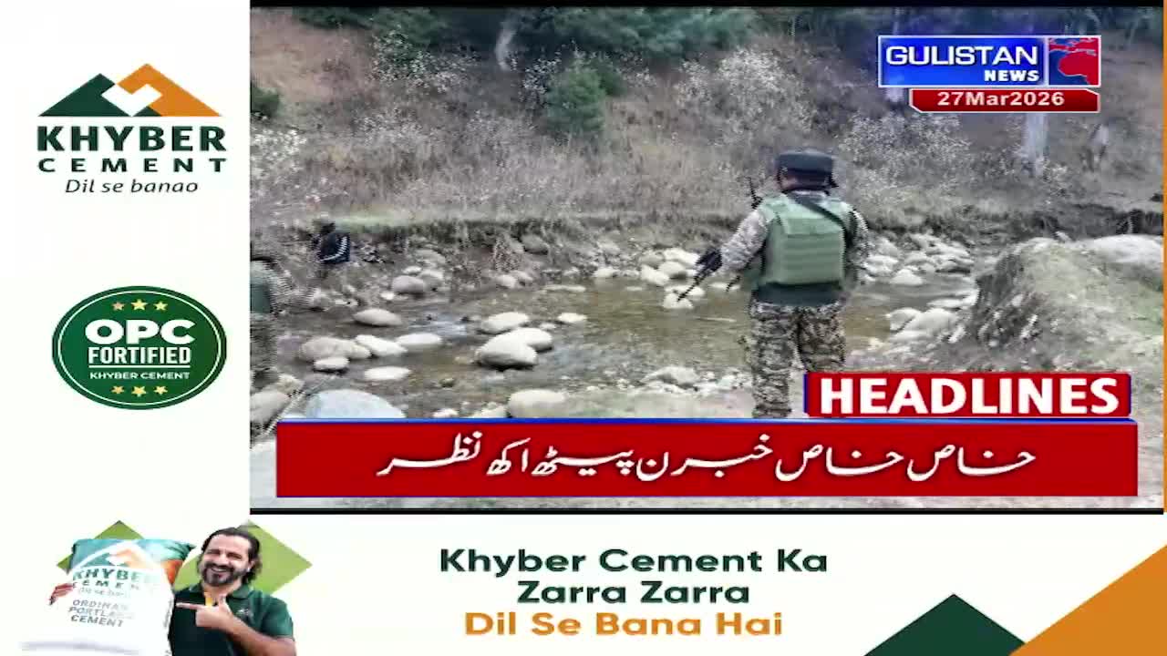 A soldier in camouflage stands guard by a shallow, rocky stream. Another figure, also in dark clothing, moves further back along the bank.