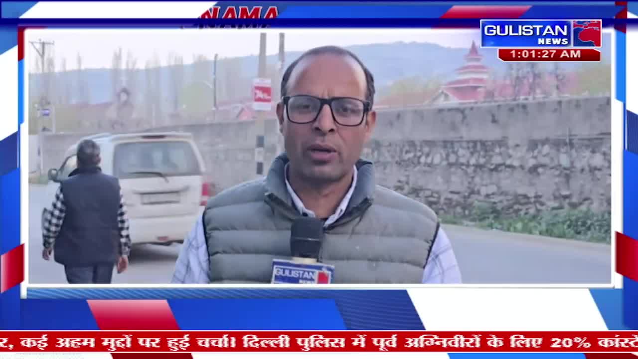 A man in a vest and glasses speaks into a microphone, reporting for Gulistan News. Behind him, a white van drives past on a road lined with trees and a stone wall.