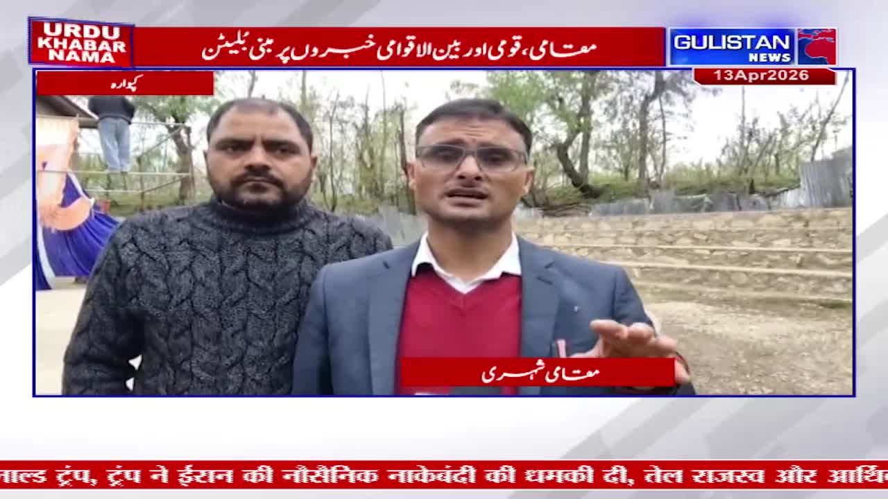 A man in a dark sweater and another in a suit and glasses stand facing the camera. The man in the suit gestures with his right hand as he speaks.